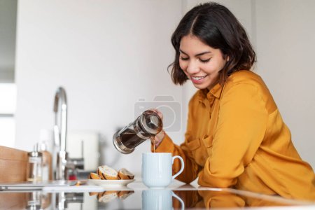 Téléchargez les photos : Closeup Shot Of Young Smiling Arab Woman Pouring Morning Coffee To Cup, Beautiful Happy Middle Eastern Female Holding French Press And Preparing Caffeine Drink White Resting In Modern Kitchen At Home - en image libre de droit