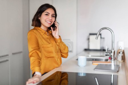 Photo for Portrait Of Smiling Young Arab Woman Talking On Cellphone In Kitchen Interior, Cheerful Millennial Middle Eastern Female Having Pleasant Phone Conversation, Smiling And Looking Away, Copy Space - Royalty Free Image