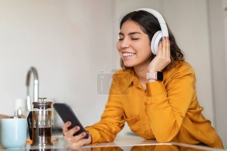 Foto de Cheerful Young Arab Female With Smartphone And Headphones Relaxing In Kitchen, Happy Smiling Middle Eastern Woman Listening Music Online On Mobile Phone, Enjoying Favorite Playlist, Closeup - Imagen libre de derechos