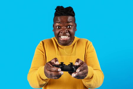 Téléchargez les photos : Excited black guy playing video games with joystick over blue background, happy african american male gamer looking at camera with excitement while standing isolated over blue background, copy space - en image libre de droit