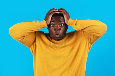 Téléchargez les photos : Portrait Of Shocked Black Young Man Opening Mouth And Touching Head, Surprised Millennial African American Guy Standing Isolated Over Blue Studio Background, Emotionally Reacting To News - en image libre de droit