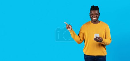 Foto de Young Black Man Holding Smartphone And Pointing Aside At Copy Space With Finger, Smiling Millennial African American Guy Recommending Nice Offer While Standing Isolated Over Blue Background, Panorama - Imagen libre de derechos