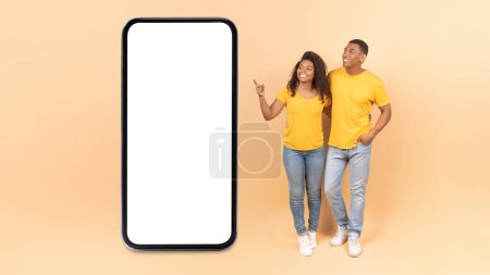 Téléchargez les photos : Happy african american spouses embracing near large smartphone, lady pointing at blank screen, advertising mobile app or offer, yellow studio background. Mockup, full length shot - en image libre de droit