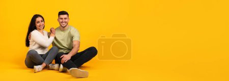 Photo for Smiling millennial arabic female and male in casual sit on floor with free space isolated on orange background, studio, panorama. Couple relaxing, rest in spare time, lad and offer, love, relationship - Royalty Free Image