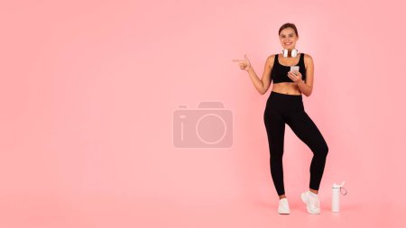 Téléchargez les photos : Fitness Ad. Smiling Sporty Woman Pointing Aside At Copy Space Over Pink Background, Athletic Young Female In Activewear With Smartphone In Hands Demonstrating Free Place For Advertisement, Panorama - en image libre de droit
