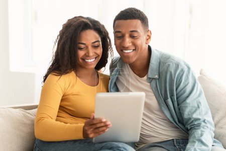 Photo for Cheerful young african american family look at tablet, make video call, surfing in internet, chatting online, sit on sofa in living room interior. New normal at home, social distancing and new app - Royalty Free Image