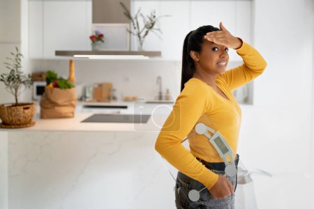 Foto de Frustrated confused young african american woman with scales, take hand to forehead in modern kitchen interior with paper package with grocery. Slimming, overweight, diet and sport, body, health care - Imagen libre de derechos