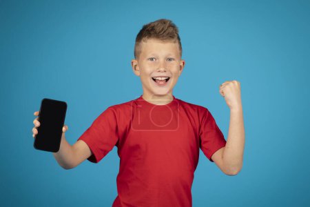 Téléchargez les photos : Excited preteen boy demonstrating smartphone with blank black screen at camera while standing isolated over blue background, cheerful male kid showing copy space for mobile advertisement, mockup - en image libre de droit