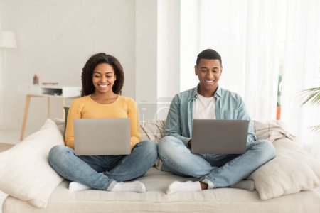 Photo for Cheerful young african american guy and lady with laptops work, surfing in internet, sit on sofa in minimalist living room interior. New normal, business, freelance and free time with device at home - Royalty Free Image