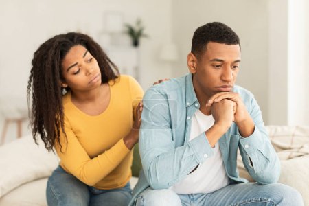 Téléchargez les photos : Serious young african american female calms offended sad male sit on sofa in room interior. Reconciliation after quarrel, relationship problems, bad news reaction and crisis, support, care at home - en image libre de droit