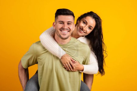 Foto de Smiling handsome millennial strong arab guy hold lady on back, have fun together, isolated on orange background, copy space, studio, close up. Couple enjoy free time, entertainment, love and romance - Imagen libre de derechos