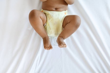 Photo for Cropped Shot Of Little African American Baby Wearing Reusable Diaper Lying On Bed, Bottom Part Of Unrecognizable Black Infant Boy Or Girl Resting On White Bedsheet In Bedroom, Top View - Royalty Free Image