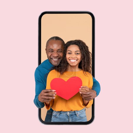 Foto de Love and care. Happy black spouses hugging holding red paper card heart standing inside cellphone screen, colorful studio background, smiling man embracing young wife. St. Valentines Day - Imagen libre de derechos