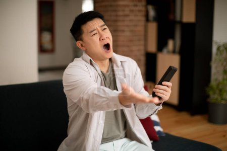 Photo for Mature korean man emotionally reacting to defeat of sports team or shocking news on television, sitting on sofa at home. Asian male expressing disbelief or indignation indoors - Royalty Free Image