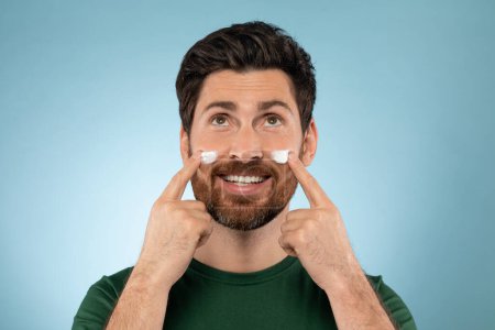 Téléchargez les photos : Bearded handsome man applying face cream on cheeks, standing on blue studio background, looking and smiling at camera. Good looking guy using beauty products for his face - en image libre de droit