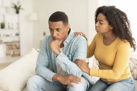 Téléchargez les photos : Apologize at home. Sad young african american female calms offended guy, support and caring, sit on sofa in room interior. Sorry after quarrel, relationship problems, bad news reaction and crisis - en image libre de droit