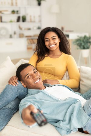 Téléchargez les photos : Glad young african american guy and lady rest and relax together in free time, watching tv with remote control on sofa in living room interior, vertical. Weekend, movie evening, entertainment at home - en image libre de droit
