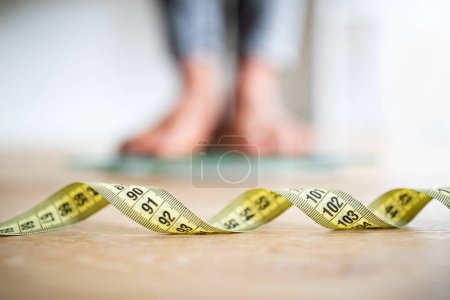 Téléchargez les photos : Feet of young african american woman on floor with selective focus on measuring tape in room interior, blurred, close up. Slimming, weight loss and overweight, diet and sport, body and health care - en image libre de droit