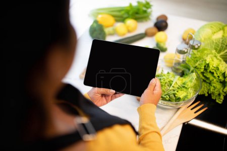 Photo for Young african american chef lady in apron cuts salad, use tablet with blank screen at table with vegetables in kitchen interior, cropped. Food blog, lesson of cook healthy food at home, ad and offer - Royalty Free Image