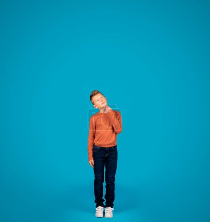 Photo for Thoughtful Preteen Boy Looking Up While Standing Over Blue Studio Background, Pensive Caucasian Male Child Touching Chin, Thinking And Considering Options, Full Length Shot, Copy Space - Royalty Free Image