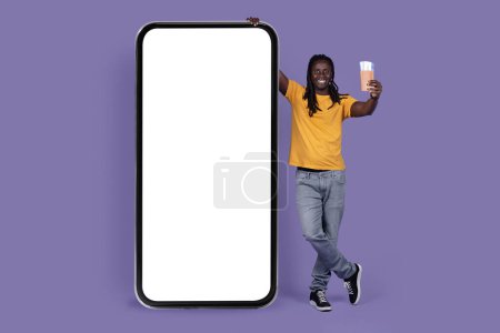 Téléchargez les photos : Happy handsome stylish long-haired young african american guy posing by huge cell phone with white blank screen for mockup, showing passport on purple, booking hotel and tickets online, full length - en image libre de droit
