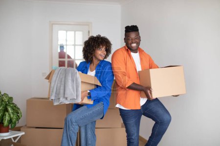 Foto de Moving Concept. Portrait Of Cheerful Young Black Couple With Cardboard Boxes In Hands Standing In Living Room Interior, Happy African American Spouses Carrying Belongings, Relocating To New Flat - Imagen libre de derechos