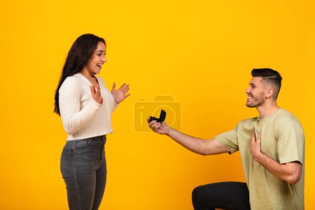 Téléchargez les photos : Smiling millennial arab guy kneeling giving ring box to happy excited bride, isolated on orange background, studio. Surprise, marriage proposal, engagement, love and romance, relationship anniversary - en image libre de droit