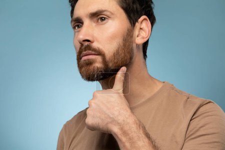 Photo for Portrait of handsome caucasian bearded man examining his face, touching beard while posing over blue background, studio shot, closeup shot - Royalty Free Image