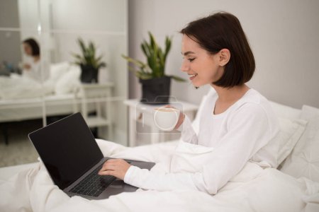 Photo for Positive pretty brunette young woman using laptop with black empty screen while staying in bed at weekend at home, typing on notebook keyboard and smiling, side view, copy space, mockup - Royalty Free Image