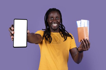 Téléchargez les photos : Cheerful happy handsome young black guy with dreadlocks in yellow t-shirt tourist showing phone with white blank screen, passport and flight tickets, showing travelling mobile app, purple background - en image libre de droit