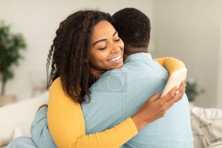 Téléchargez les photos : Cheerful millennial african american lady hugging man and chatting in smartphone, ordering goods in home interior. Addiction from device and social networks, problems in relationships, ad and offer - en image libre de droit
