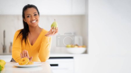 Foto de Cheerful young african american female recommending pear and refusing donut at table in modern light kitchen interior with copy space, panorama, blurred. Diet, health care, sweets and weight loss - Imagen libre de derechos