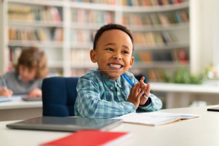 Photo for Happy african american primary school boy sitting at desk in classroom, writing in notebook and smiling, free space. Group of diverse classmates studying in the background - Royalty Free Image