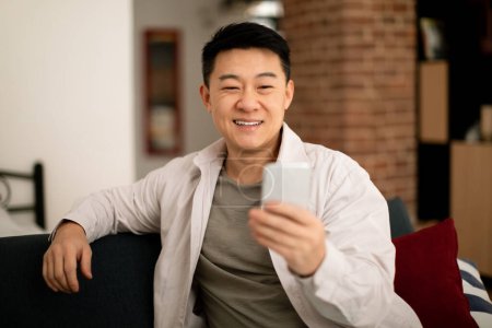 Foto de Happy mature asian man in casual watching video or chatting on smartphone, resting on sofa in living room interior. Business and work remotely at home, blog, new normal and new app - Imagen libre de derechos