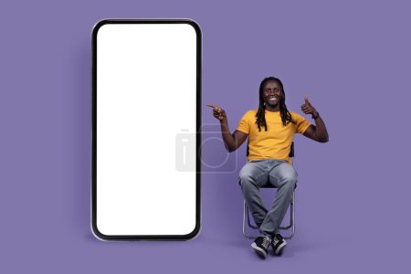 Téléchargez les photos : Positive handsome stylish african american young man with long dreadlocks sitting on chair by huge smartphone with white blank screen, showing thumb up, mockup, isolated on purple studio background - en image libre de droit