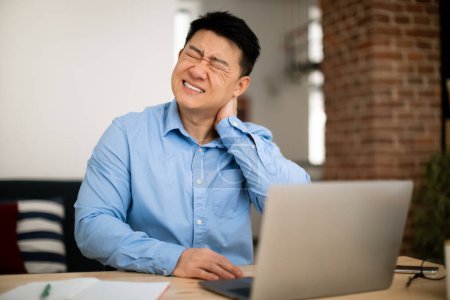 Photo for Neck ache. Mature asian man massaging aching neck, suffering from pain while working on laptop at table at home office. Poor posture issue and arthritis problem - Royalty Free Image
