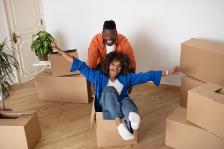 Téléchargez les photos : Cheerful Young Black Spouses Having Fun Together While Moving To New Apartment, Happy Excited African American Woman Riding In Cardboard Box In Living Room, Positive Couple Fooling At Home - en image libre de droit