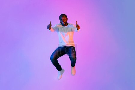 Téléchargez les photos : Cool happy handsome mature african american man in casual outfit jumping up and showing thumb ups on colorful neon studio background, copy space for advertisement, full length - en image libre de droit