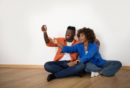 Foto de Happy Young Black Spouses Sitting On Floor And Holding Home Keys, Smiling African American Couple Celebrating Moving To Their New Apartment After Buying Or Renting Property, Copy Space - Imagen libre de derechos