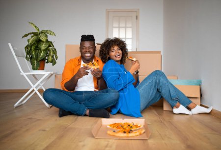 Téléchargez les photos : Young black couple sitting on floor and eating pizza, resting after moving home, happy african american spouses relaxing in living room among packed cardboard boxes, celebrating relocation day - en image libre de droit