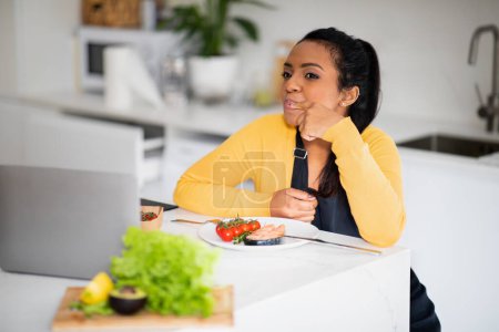 Photo for Smiling hangry young african american female in apron licks finger, looks at laptop and fresh fish, enjoys dish in modern kitchen interior. Delicious diet, cook healthy food at home, dinner with blog - Royalty Free Image