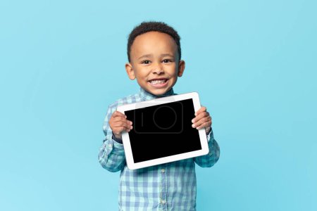 Foto de Cute african american kid boy holding digital tablet with blank black screen, recommending new educational app or website, standing over blue studio background, mockup - Imagen libre de derechos