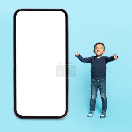 Photo for Cute african american child boy in casual outfit standing by big smartphone with white blank screen and showing thumbs up, recommending nice app, blue studio background - Royalty Free Image