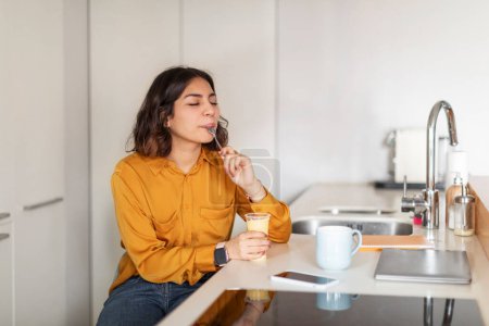 Téléchargez les photos : Young Arab Woman Sitting At Kitchen Counter And Eating Tasty Pudding From Plastic Cup, Happy Millennial Middle Eastern Female Licking Spoon While Enjoying Delicious Dessert Meal At Home, Copy Space - en image libre de droit