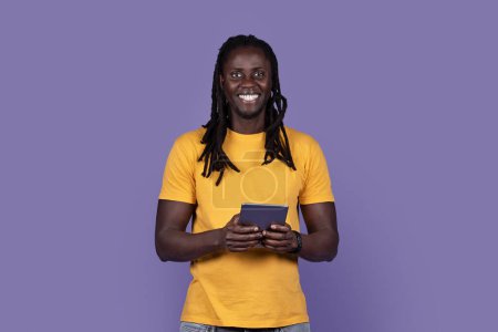 Foto de Happy smiling stylish long-haired young black guy in casual outfit using modern digital pad on purple studio background, checking newest entertaining mobile application, copy space - Imagen libre de derechos