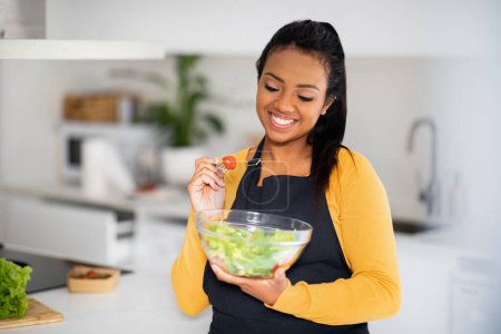 Foto de Smiling young african american female chef in apron eating organic vegetable salad, enjoy fresh dish in modern kitchen interior. Delicious diet, lose weight and cooking healthy homemade food at home - Imagen libre de derechos