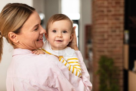 Photo for Maternal love. Mother holding cute infant girl on hands and cudding, embracing baby and smiling, standing in bedroom interior, free space - Royalty Free Image