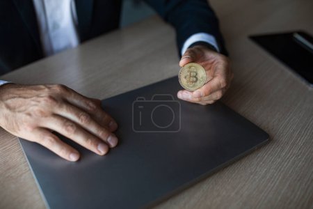 Foto de Businessman in suit holding bitcoin in his hand, sitting at workplace in office interior, cropped. Professional planning budget, successful business and savings, modern money, cryptocurrencies - Imagen libre de derechos