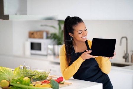 Foto de Happy young african american woman in apron show tablet with empty screen at table with fresh vegetables in modern kitchen interior. App recommendation, diet, healthy food cooking at home, blog lesson - Imagen libre de derechos