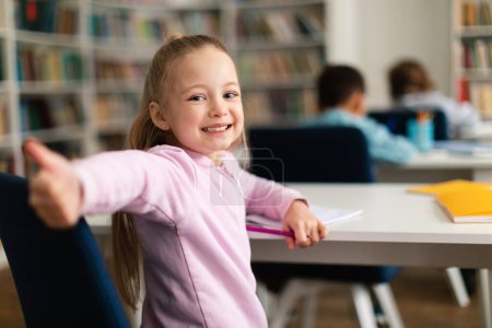Téléchargez les photos : Happy caucasian schoolgirl gesturing thumbs up and smiling to camera, sitting at school in classroom with multiracial kids, free space. Education concept - en image libre de droit